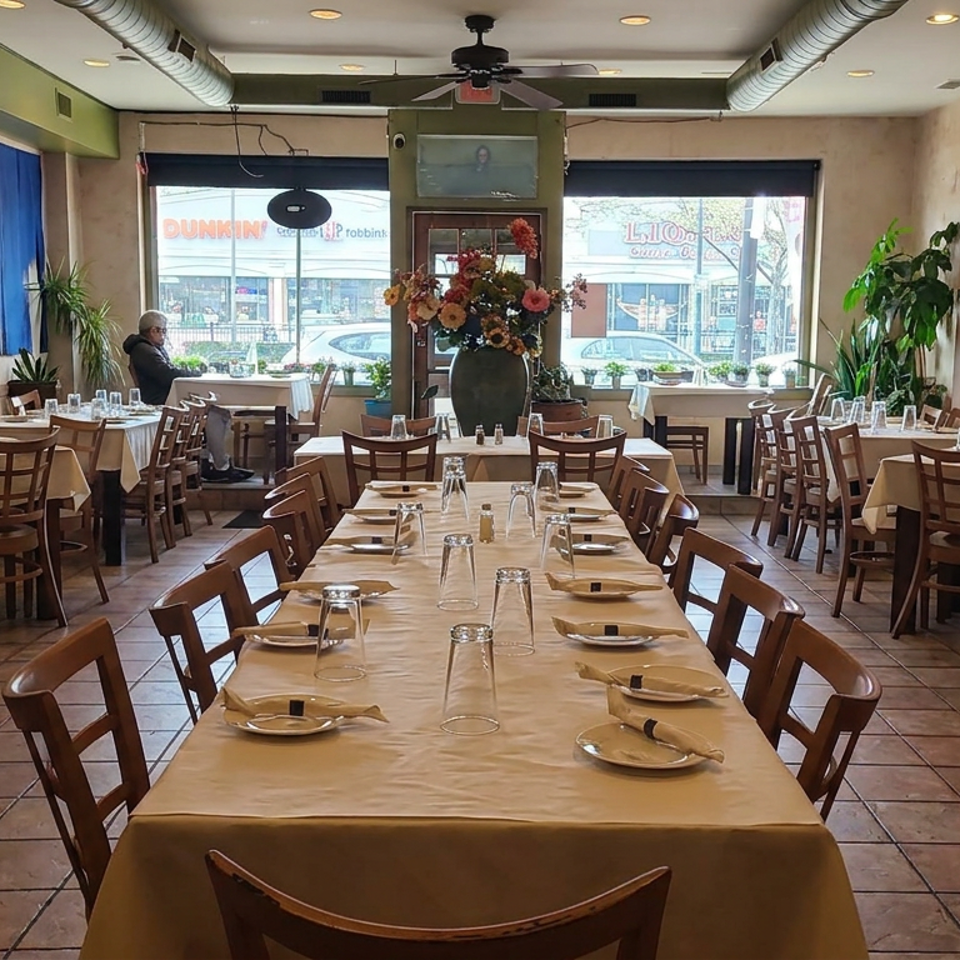 Bright restaurant dining room with wood tables, cream tablecloths, place settings, potted plants, and large street-facing windows.