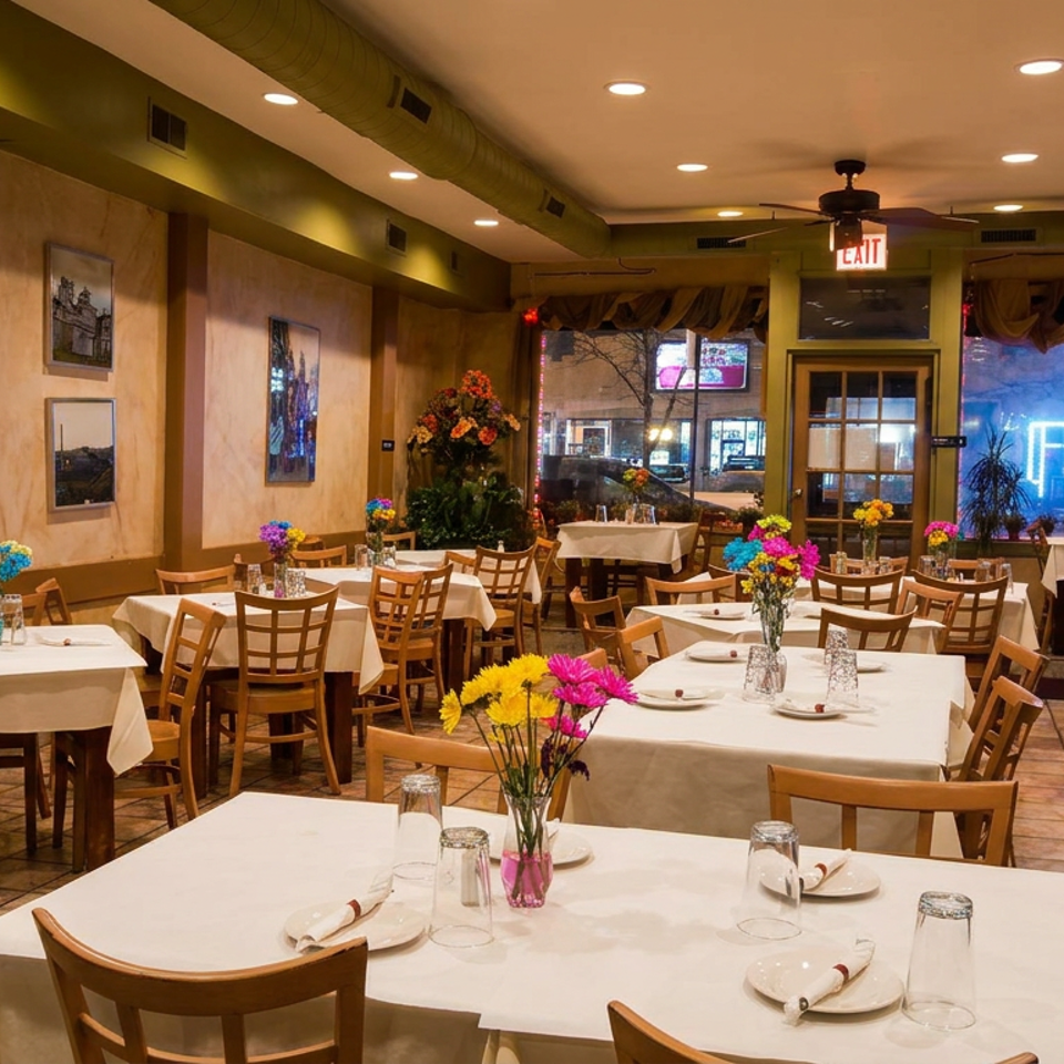 Warmly lit restaurant dining room with white tablecloths, wooden chairs, colorful flower centerpieces, framed artwork, and street-facing windows.